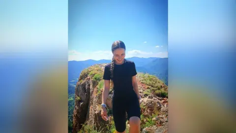 Smiling young woman in black athletic wear standing on a rocky mountain peak under a clear blue sky.