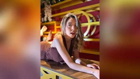 Young woman in a brown sleeveless top leaning on a wooden table in a warm, moody indoor setting.