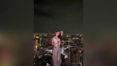 CheCascano poses on a balcony at night overlooking a bright city skyline, wearing a white tank top and patterned pants.