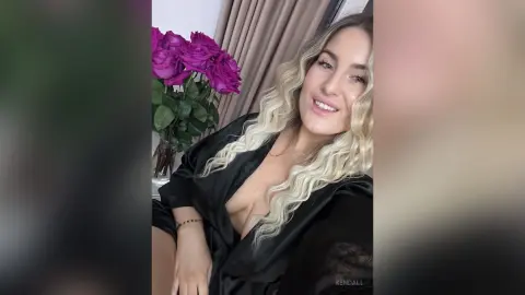 Smiling blonde woman in a black satin robe relaxing near a bouquet of purple roses in a bright room.