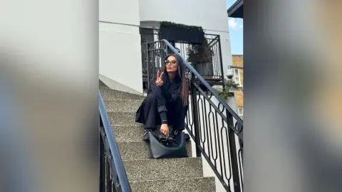 Stylish woman in a black outfit and sunglasses sitting on outdoor stairs with a chic black handbag.