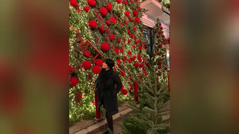 Woman in a black winter coat looking up at a festive wall of red Christmas ornaments and twinkling lights.