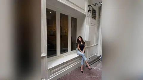 OliviaHardies sits on a white window ledge outside a building, wearing a black tank top and light blue jeans.