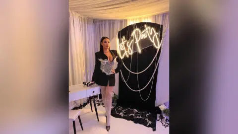 AnnaKarev poses in a black blazer and sheer lace leggings in front of a black backdrop with a neon sign.