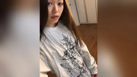 Young woman in a white graphic long-sleeve shirt posing in a cozy indoor setting.