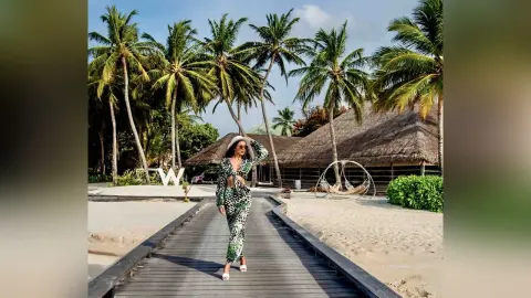 Stylish woman in a green patterned jumpsuit walking on a wooden boardwalk at a tropical beach resort.