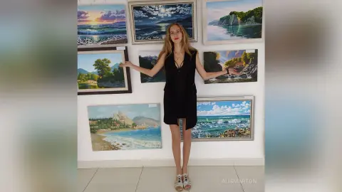 Blonde woman in a black dress standing in an art gallery surrounded by coastal landscape paintings.