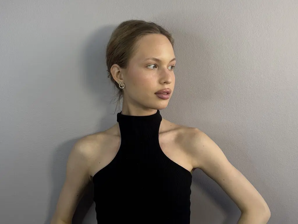 MalliaLucioni looks away from the camera, wearing a black sleeveless turtleneck top with her hair pulled back against a plain grey wall.
