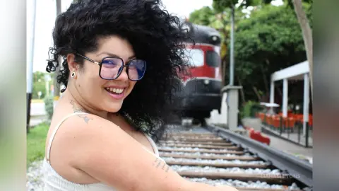 Smiling woman with curly hair and glasses in a white tank top sitting by train tracks on a sunny day.