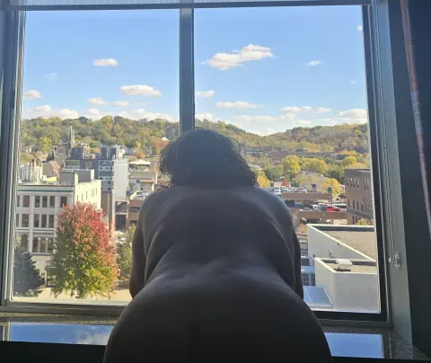 Nude woman sitting on a windowsill looking out at a sunny autumn cityscape through a large glass window.