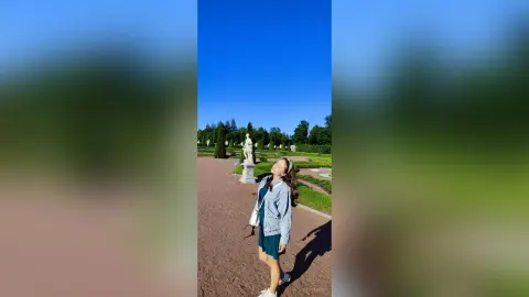 Cheerful young woman in a denim jacket and dark dress enjoying a sunny day at a classical garden park.