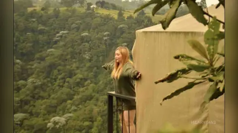 Blonde woman in a green sweater leaning on a balcony overlooking a lush, misty forest landscape.