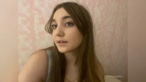 MartineKibbey looks at the camera with long brown hair and a grey tank top against a pink patterned wall.
