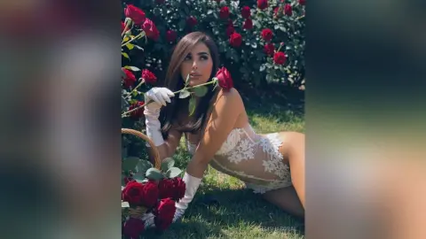 AllishaCoral — Elegant woman in white lace lingerie and gloves holding a red rose in a lush rose garden.