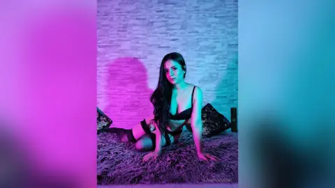 Woman in black lingerie sitting on a shag rug against a textured wall with moody pink and blue neon lighting.