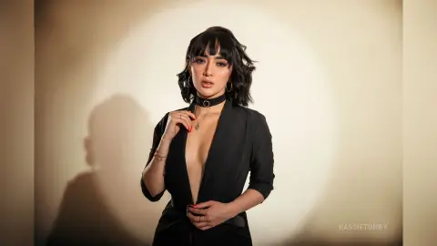 Elegant woman in a black plunging jumpsuit and choker posing against a warm studio backdrop.