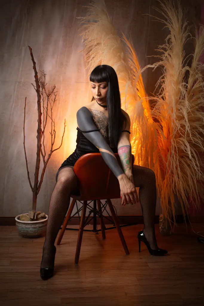 SamMansson wears glasses and a black lace corset while posing with her hands in her dark hair, showing off her tattoos.