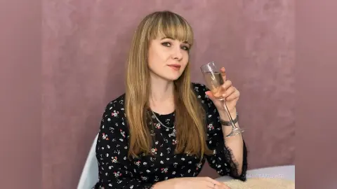 Blonde woman in a black floral blouse holding a champagne glass against a soft pink background.