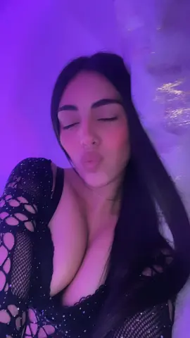Young woman with long dark hair in a black mesh top making a kissy face under purple ambient lighting.