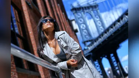 Stylish woman in sunglasses and a grey trench coat posing near the Manhattan Bridge on a sunny day.