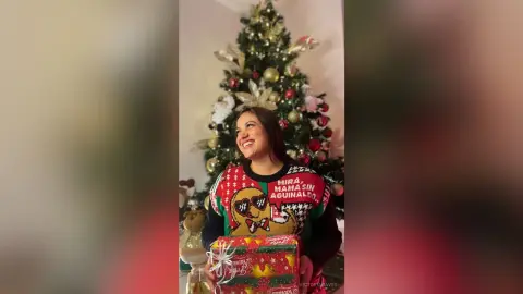 Smiling woman in a festive red Christmas sweater holding a wrapped gift near a decorated holiday tree.