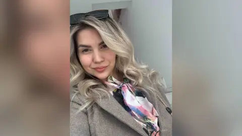 RaisaAllens smiles with her long blonde hair and sunglasses resting on her head, wearing a grey coat and a patterned silk scarf.