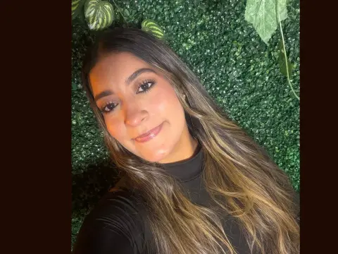 Sharlotte_Rose smiles while wearing a black turtleneck against a green leafy wall background.