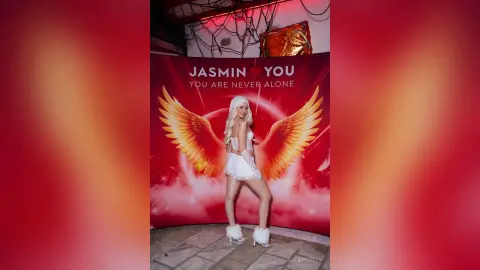 Blonde woman in white angel costume with large wings posing against a red dreamy backdrop.