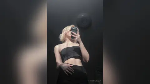 Blonde woman in a black crop top taking a moody mirror selfie against a dark minimalist wall.