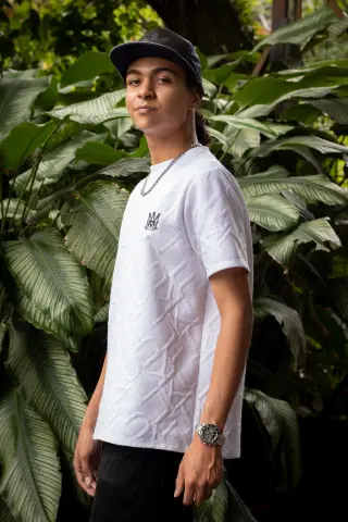 KylePresly stands outdoors against large green tropical leaves wearing a white textured t-shirt, a black cap, and a silver chain.