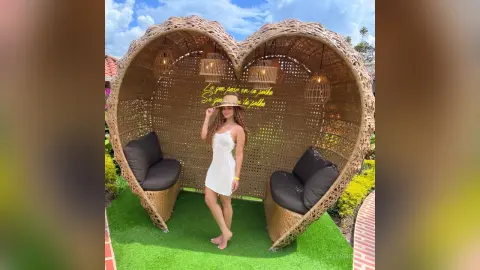 Woman in a white summer dress standing in a heart-shaped wicker lounge chair in a sunny garden setting.