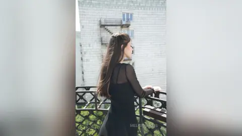 Melancholic woman in a black long-sleeve dress standing on a balcony overlooking urban buildings.