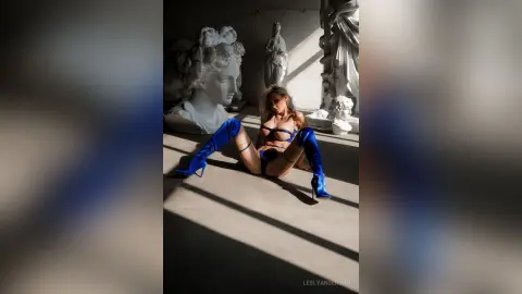 LeslyAnderson sits on a floor between marble statues wearing a black bra and blue thigh-high boots.