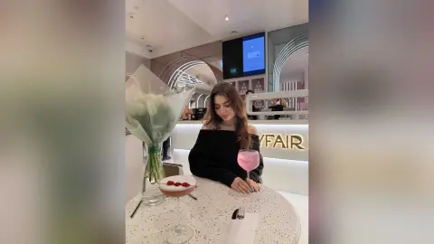 Young woman in a black off-the-shoulder top enjoying a pink cocktail at a modern cafe with fresh flowers.