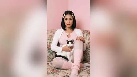Young woman in pink loungewear sitting on a floral sofa with a black and white cat in a soft pink room.