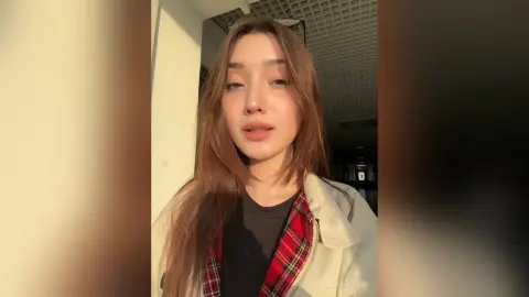 Young woman with long brown hair wearing a plaid shirt and white jacket in warm sunset lighting.