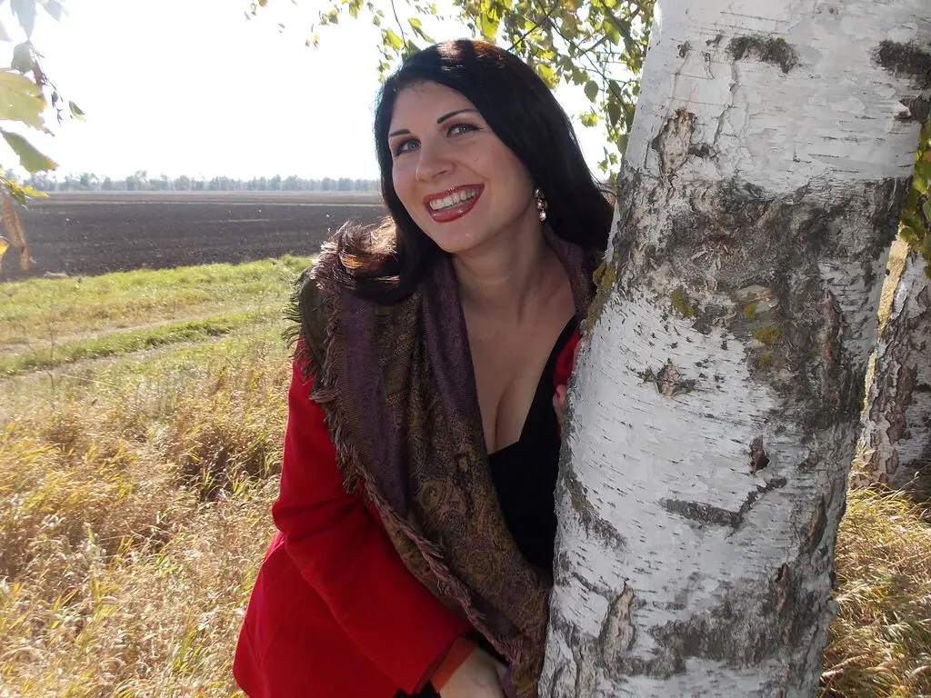 Smiling woman in a red top and patterned scarf leaning against a birch tree in a sunny autumn field.