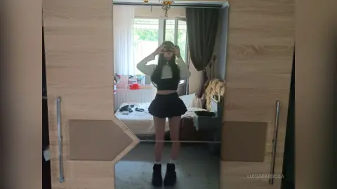 Young woman in a black crop top and mini skirt taking a mirror selfie in a bright bedroom.