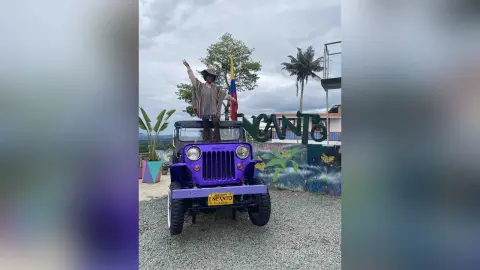 Cheerful traveler in a striped shirt and hat standing on a purple vintage jeep in a tropical outdoor setting.