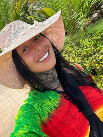 Smiling woman in a sun hat and colorful tropical shirt enjoying a sunny day in a lush garden setting.