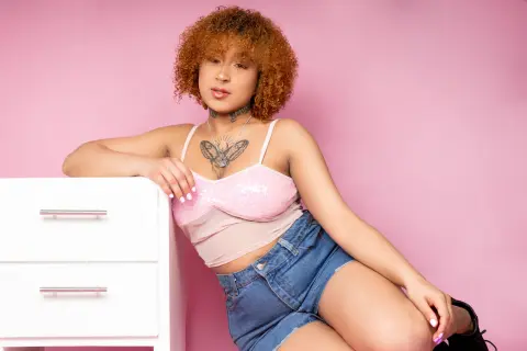 Young woman with curly hair in a pink crop top and denim shorts leaning on a white dresser against a pink background.