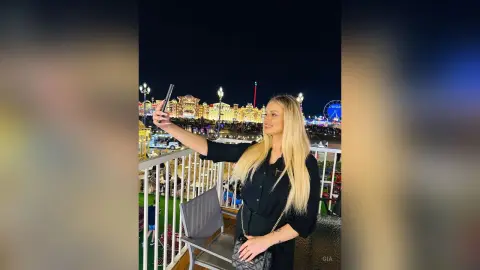 Blonde woman in a black blouse taking a selfie on a balcony overlooking a lit amusement park at night.
