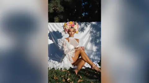 Ethereal woman in a white ruffled gown wearing a floral crown, sitting against a white backdrop in a garden.