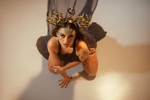 Mystical woman in black outfit wearing a golden snake crown looking up in a moody studio setting.