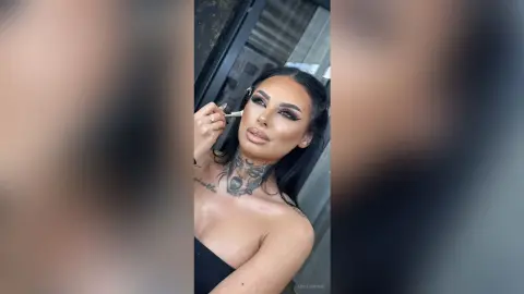 Woman with dramatic winged eyeliner and neck tattoos applying makeup in a bright indoor setting.
