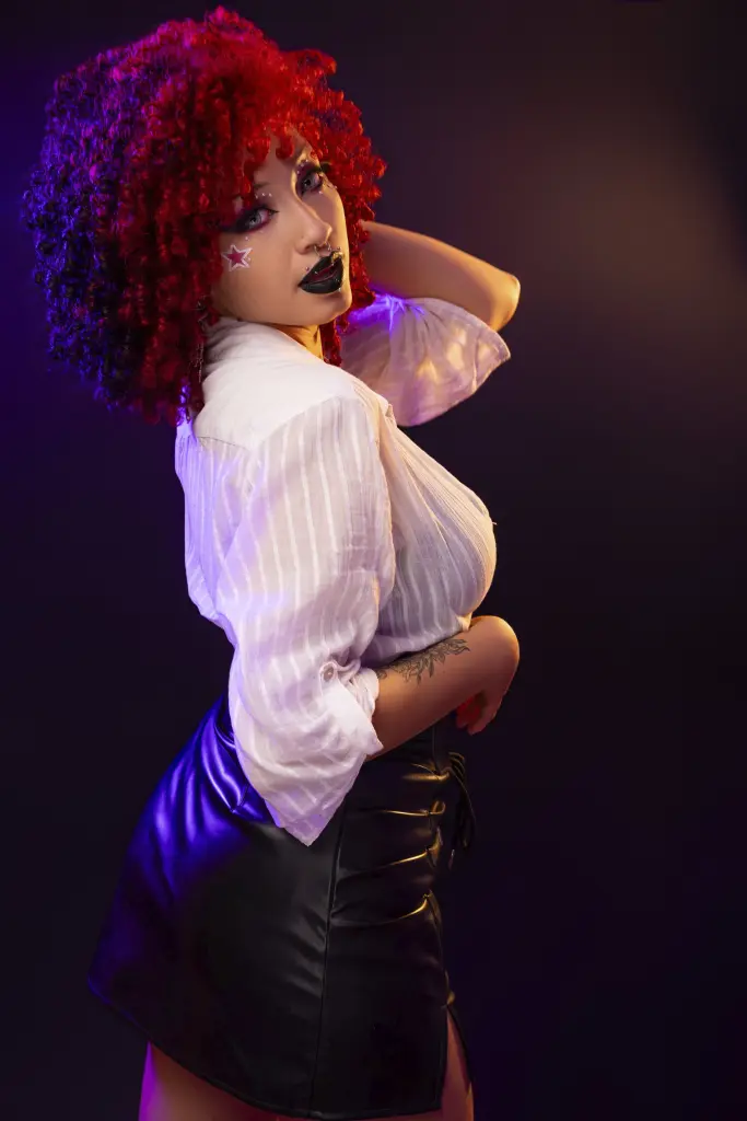 NaylaNyx poses with her red curly hair and dark lipstick, wearing an unbuttoned white shirt against a dark background.