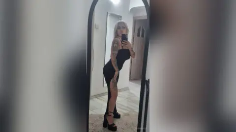 Blonde woman in a sleek black evening dress posing for a mirror selfie in a modern minimalist room.