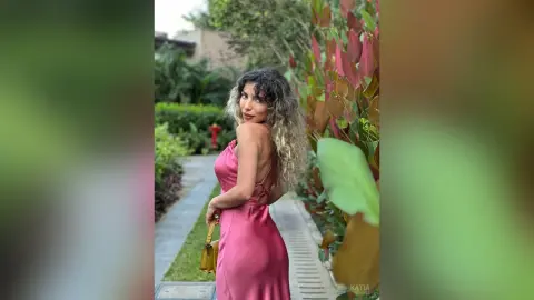 Katia poses in a pink backless dress while standing on a garden path next to tall leafy plants.