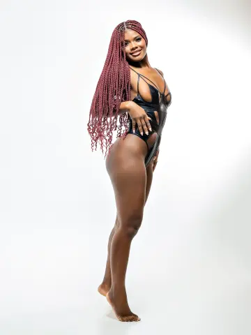 Smiling Black woman with long red braids wearing a black cutout bodysuit in a bright studio setting.