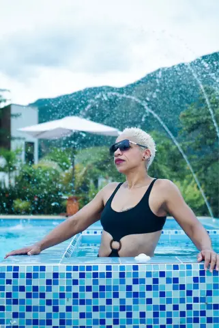 Woman in black swimsuit relaxing by a blue tiled pool with mountain views and water splashes.